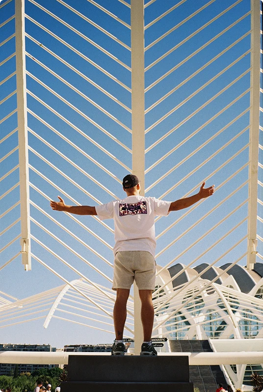 Person standing with arms outstretched under a modern white architectural structure against a clear blue sky.