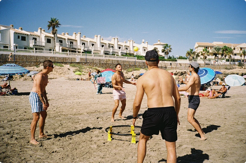 Four men playing Spikeball on a sandy beach under clear blue sky with houses and umbrellas in the background.