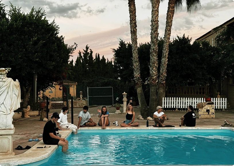 Group of six young people sitting and relaxing by a swimming pool at dusk surrounded by trees and statues.