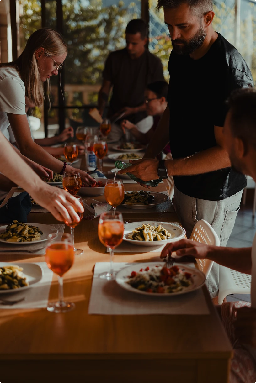 Group of people gathered around a table serving pasta dishes and pouring orange drinks into wine glasses.