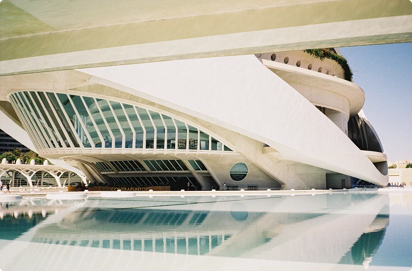 Futuristic white building with large glass windows reflecting in a calm pool of water under a clear sky.