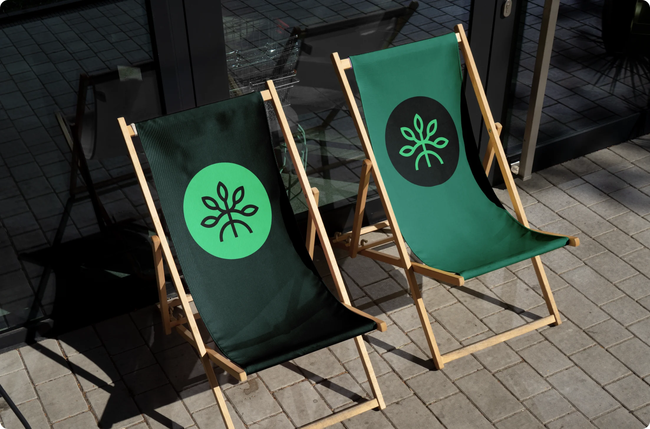 Two wooden lounge chairs with green fabric featuring a leaf logo, placed on a tiled floor outside a glass door.