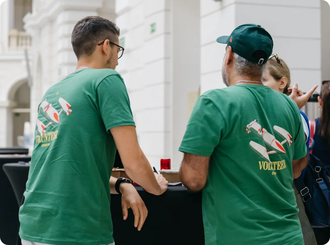 Two men in green t-shirts with airplane graphics and 'VOLUNTEER' text on the back, standing and talking indoors.