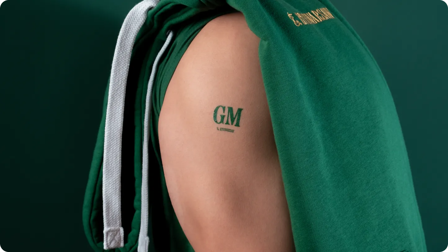 Person wearing green hoodie with sleeve pulled up, showing a small green tattoo on upper arm reading 'GM'.