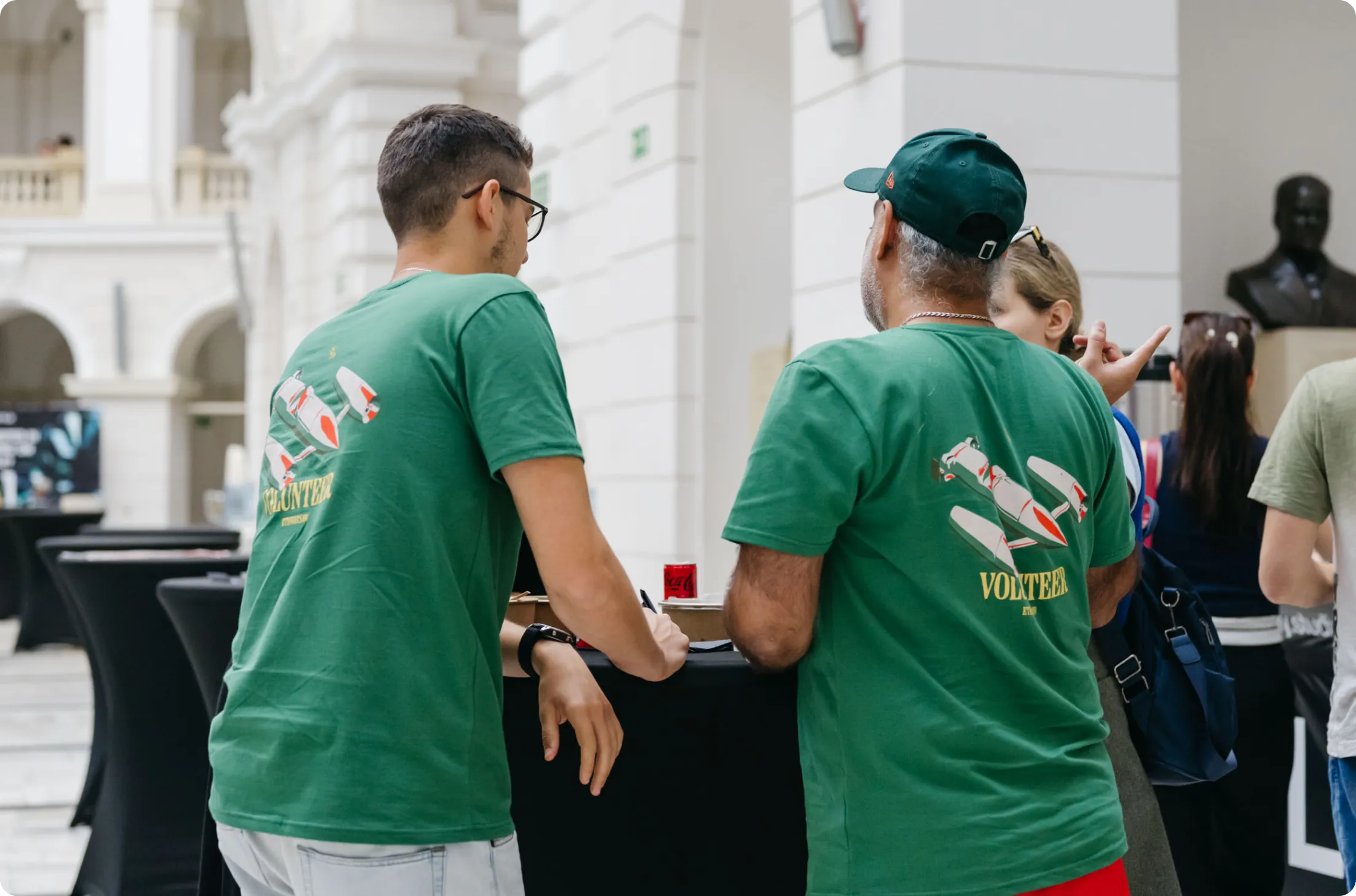 Two men wearing green volunteer t-shirts talking near a black table in a bright indoor space.