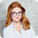 Woman with red hair and glasses wearing a white shirt against a gray background.