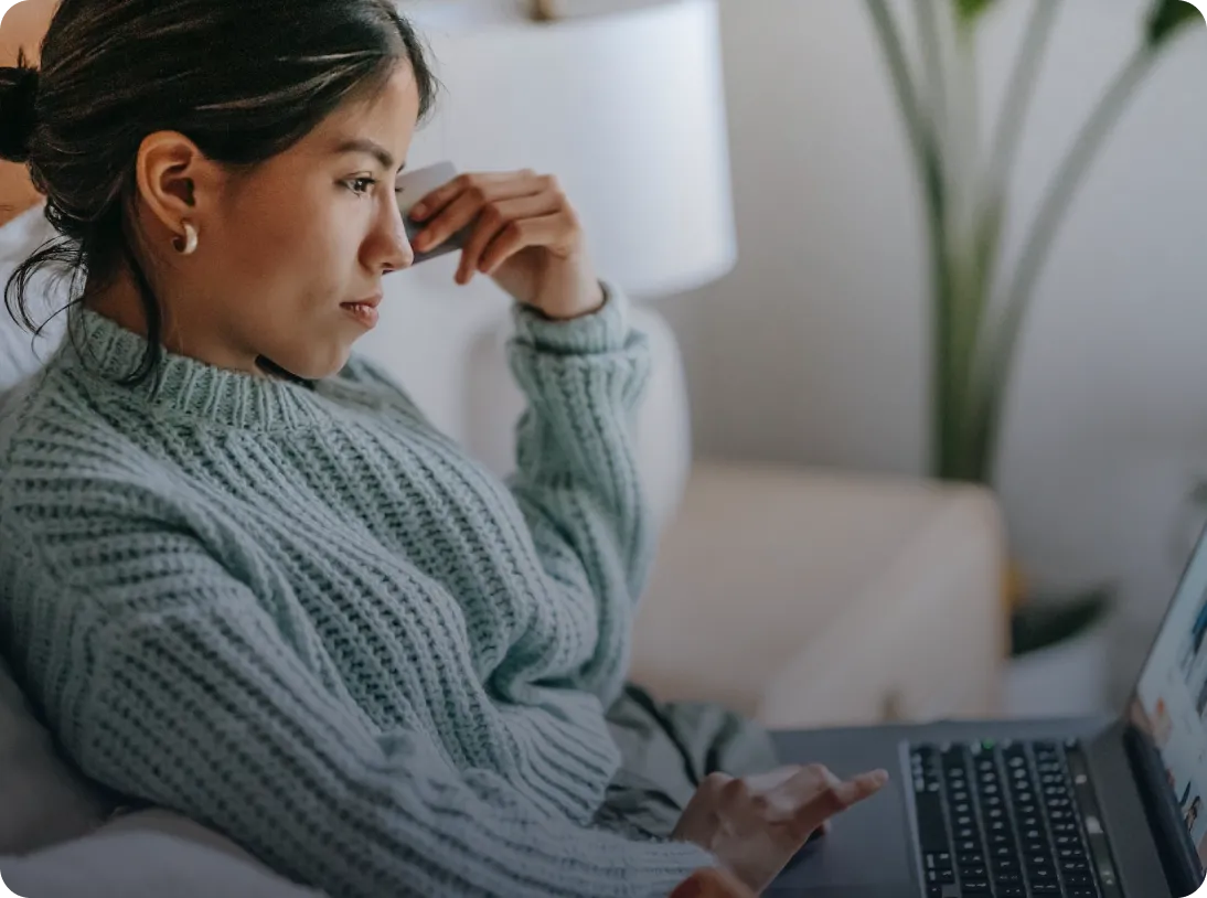 Woman in a knitted sweater sitting on a couch, holding a phone to her ear and using a laptop.