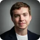 Headshot of a young man with short brown hair wearing a dark jacket and white shirt, smiling slightly against a gray background.