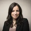 Smiling woman with long dark hair wearing a black blazer and light pink top against a neutral background.