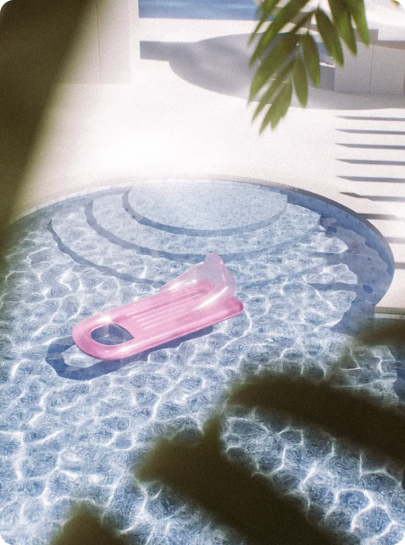 Pink inflatable pool float resting on the water in a round swimming pool with steps, partly shaded by nearby palm leaves.