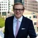 Smiling man in glasses wearing a dark suit and pink tie standing outdoors with city buildings in the background.