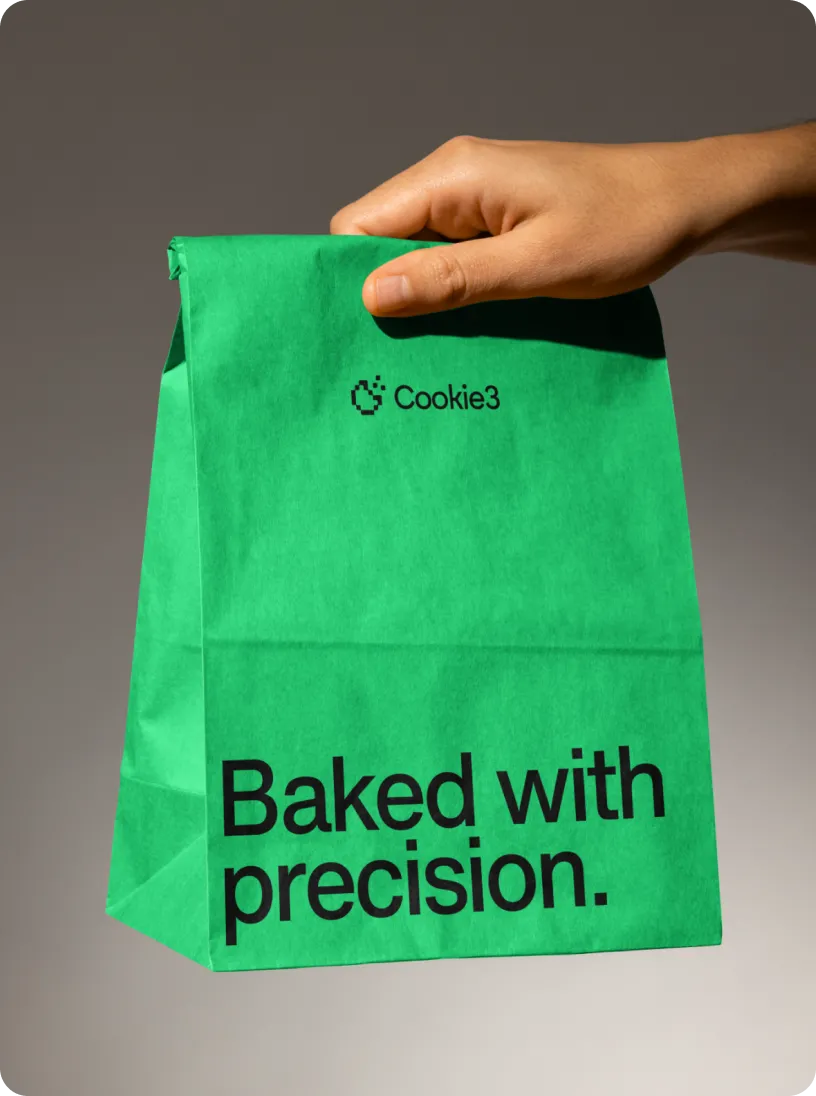 Hand holding a green paper bag with the text 'Cookie3' and 'Baked with precision.' printed on it.