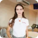 Young woman with long brown hair wearing a white polo shirt standing in a modern office space.