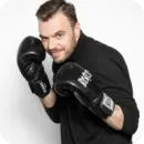Smiling man in black shirt wearing black boxing gloves in a boxing stance.