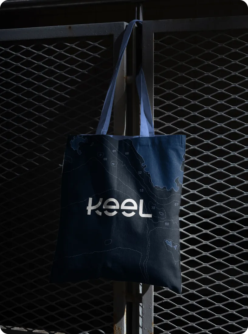 Dark blue tote bag with white topographic map lines and the word 'keel' hanging on a metal mesh gate.