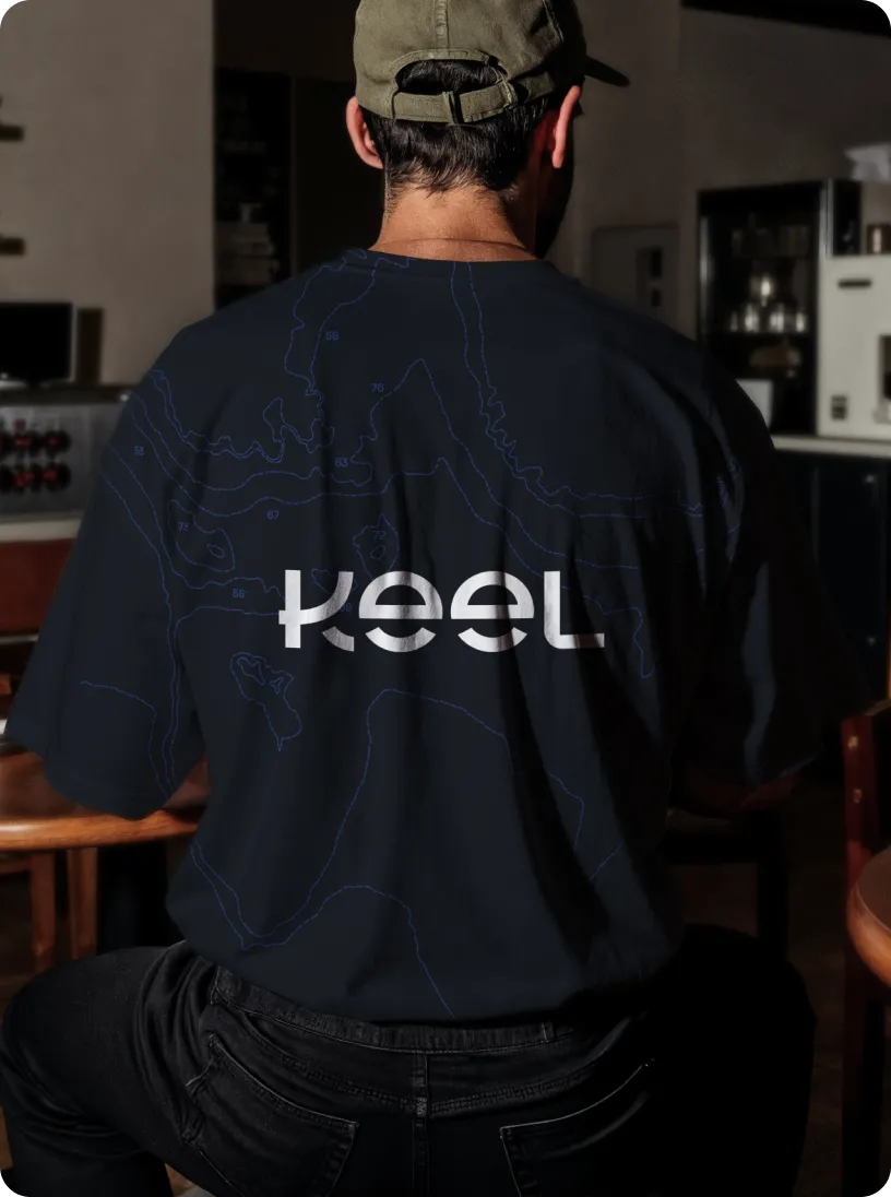 Man wearing a black t-shirt with 'keel' written on the back in reflective letters, sitting indoors, viewed from behind.