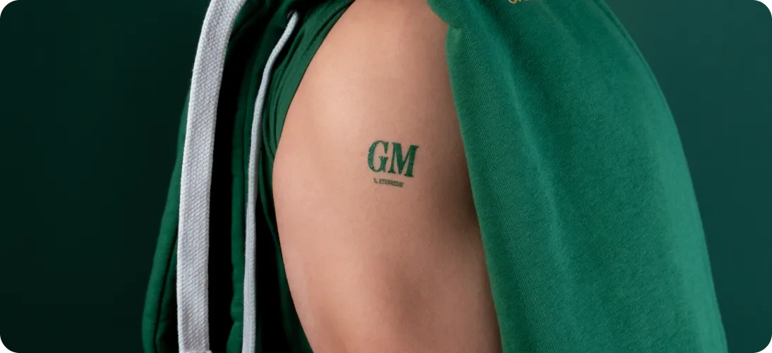 Close-up of a person's upper arm with a green tattoo reading 'GM' and smaller text beneath it, partially covered by a green hoodie.