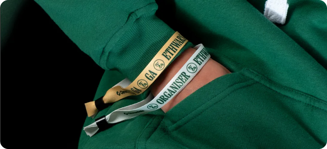 Person wearing green hoodie with two wristbands that read 'ETHWARSAW' and 'ORGANISER'.