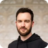 Portrait of a bearded man with dark hair wearing a black hoodie against a blurred beige background.