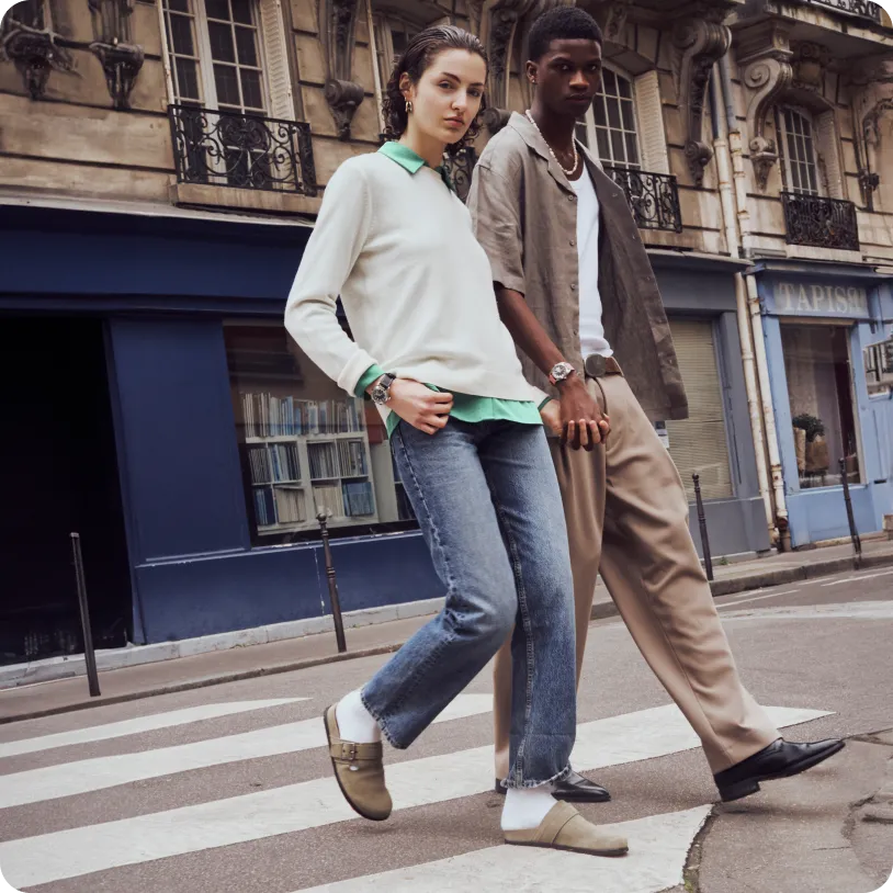 A young woman in a white sweater and jeans and a young man in beige pants and a brown shirt crossing a city street holding hands.