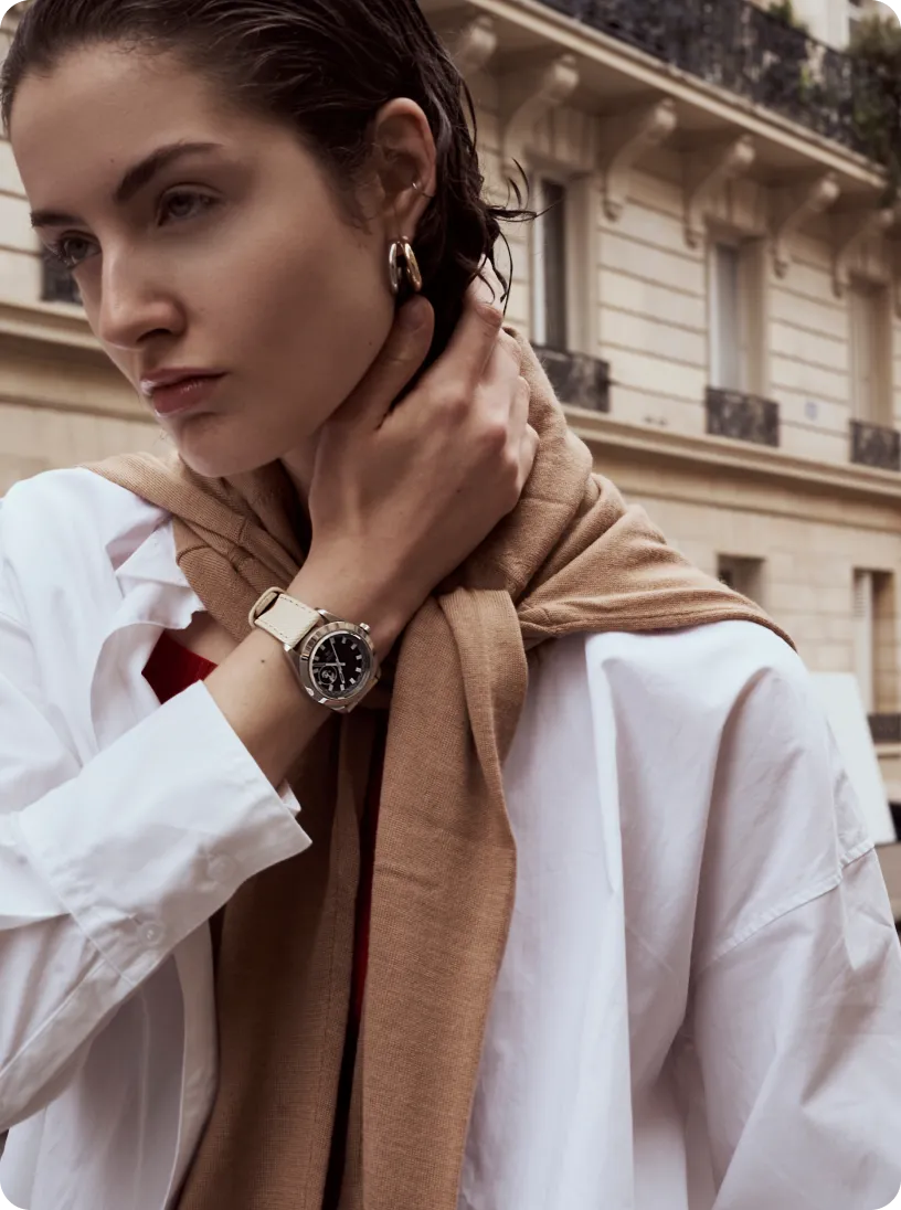 Close-up of a woman wearing a white shirt, beige scarf, and a wristwatch, touching her neck with a thoughtful expression in an urban setting.