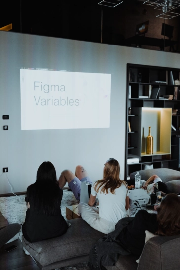 Group of people sitting in a dim room watching a projection on a wall displaying the text 'Figma Variables'.