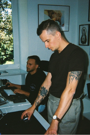 Two men working in an office, one standing with tattoos and a watch using a mouse, and the other sitting at a keyboard.