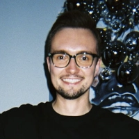 Smiling man with glasses, short dark hair, and beard wearing a black shirt, standing in front of a dark decorative background.