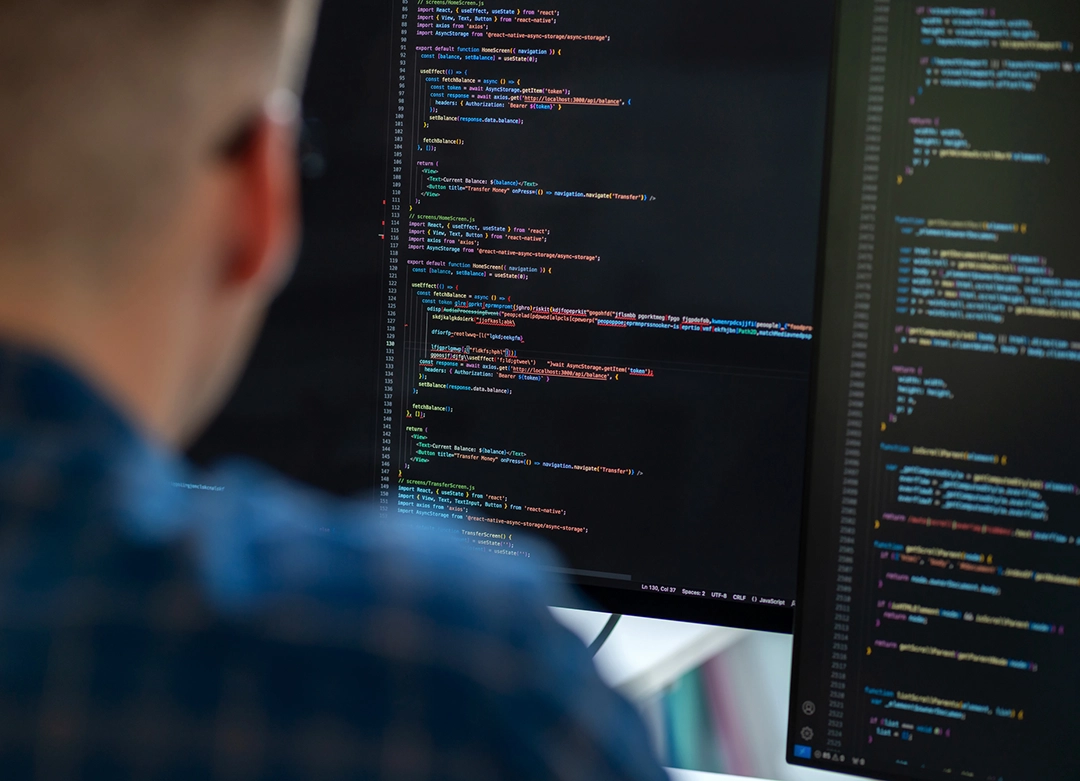 Person in glasses and checkered shirt viewing two computer monitors displaying code with a dark background.