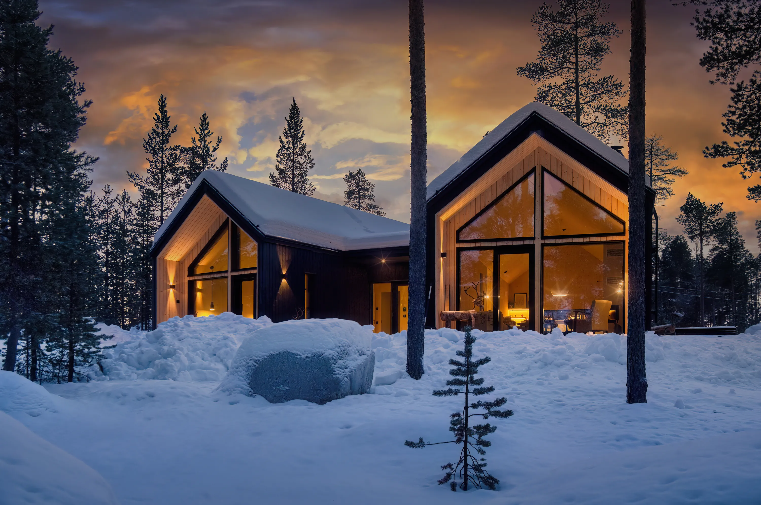 Two modern cabins in a snowy landscape at dusk, with warm interior lighting contrasting with the cool exterior.
