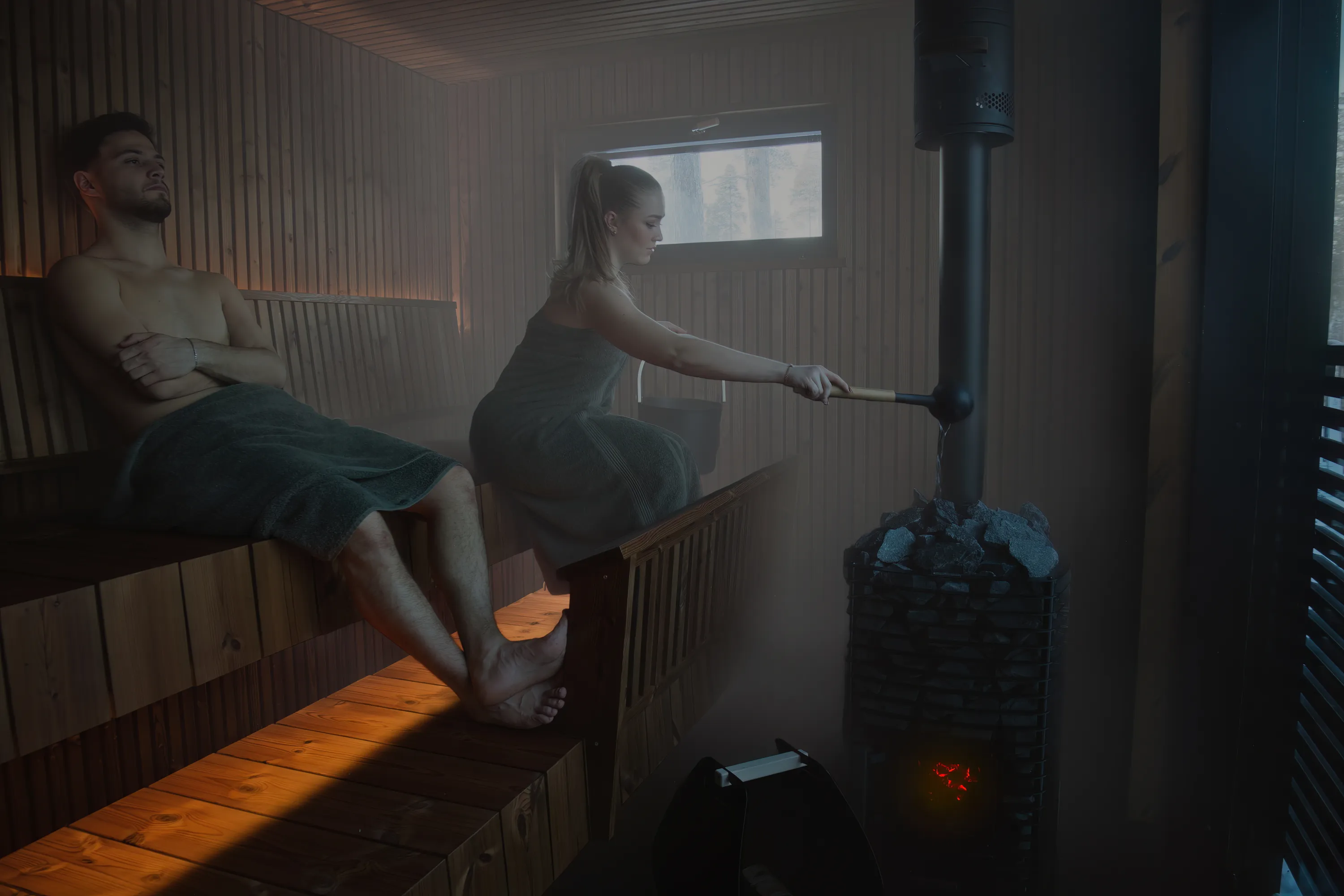 A man and a woman are in a sauna. The man is sitting on a bench with a towel wrapped around his waist. The woman is pouring water on the hot stones of a sauna stove. The sauna is made of wood and has a small window.