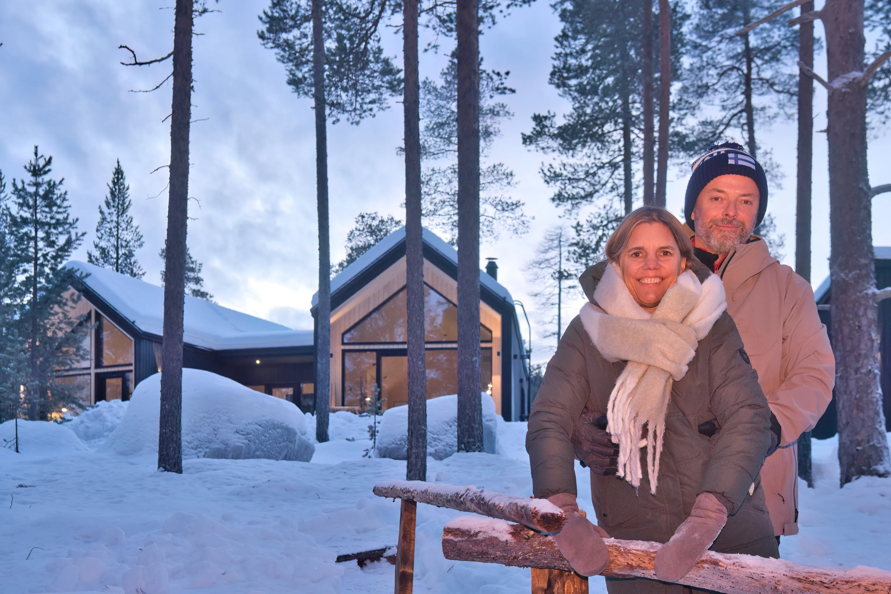 A couple stands in a snowy forest in front of a modern house, with tall trees surrounding them, under a twilight sky.