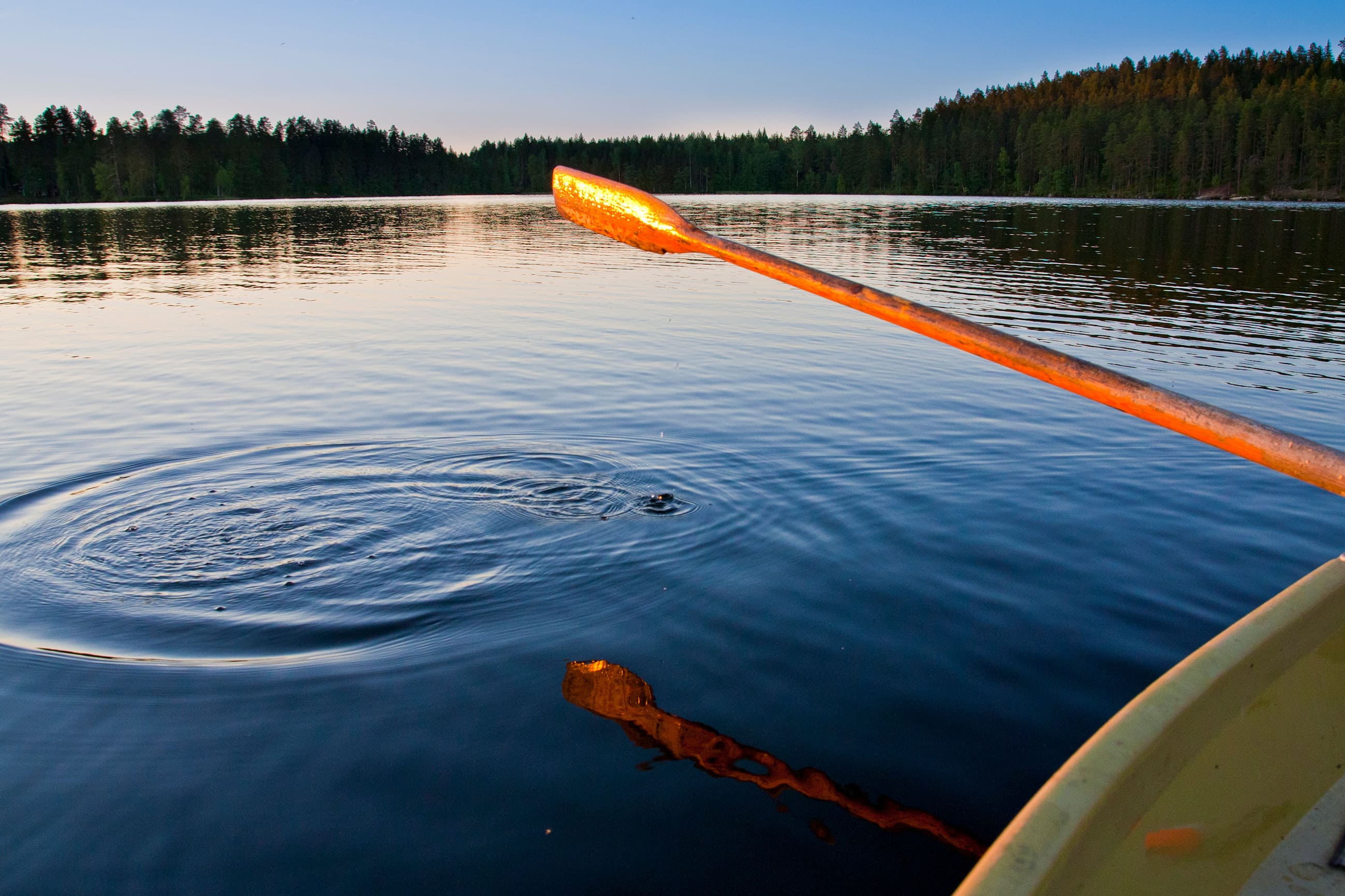 A golden paddle extends from a boat across a calm, deep blue lake at sunset. Distant dark green trees line the horizon under a pale blue sky, while concentric ripples and small bubbles spread across the water in the foreground.