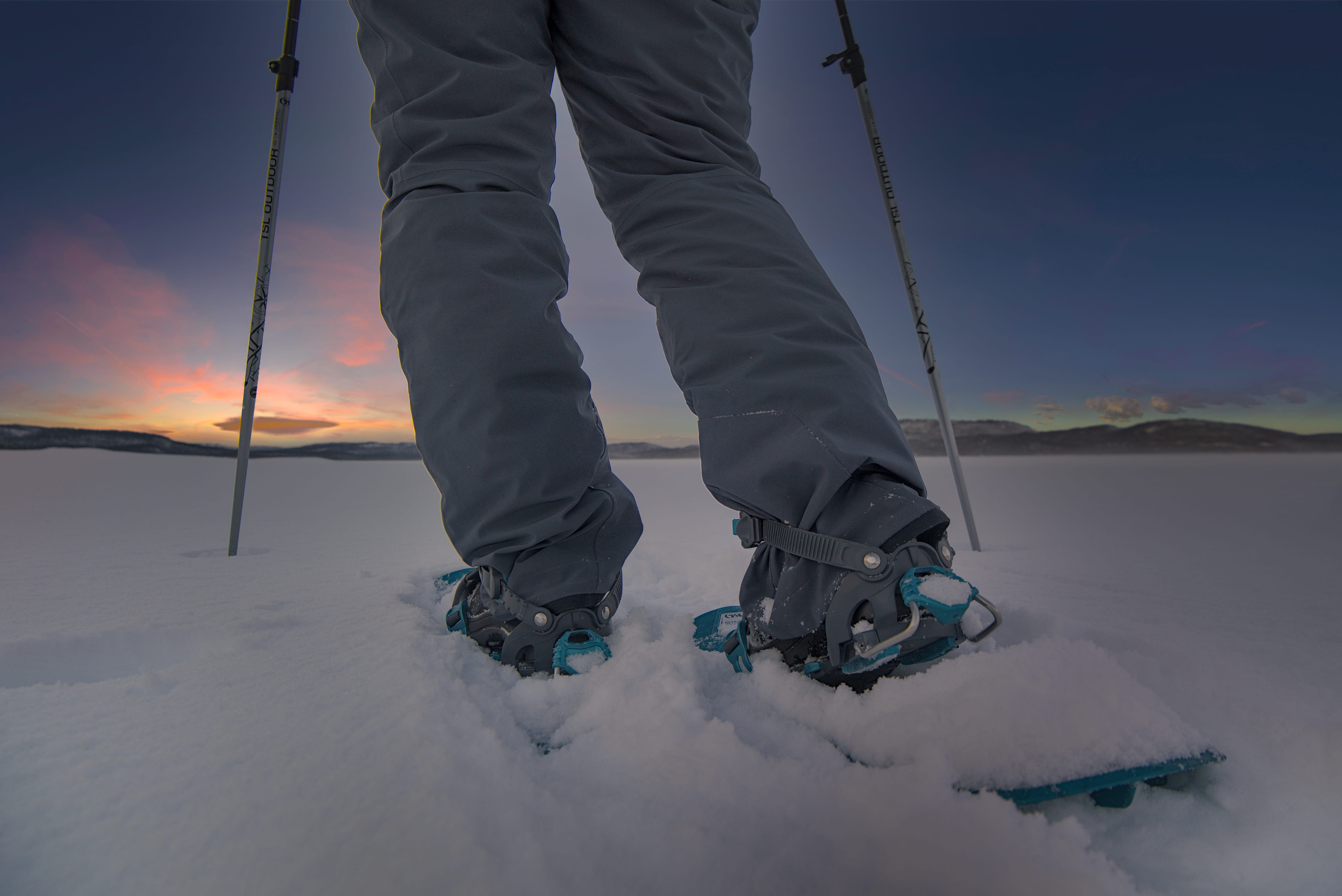 A low-angle shot captures a person's lower body, clad in dark grey snow pants and grey snowshoes with bright blue accents, standing in deep white snow. Two silver trekking poles are visible, one on each side, with 'TSL OUTDOOR' branding clearly legible on the left pole. The horizon features dark mountain silhouettes under a dramatic sky, transitioning from deep blue overhead to vibrant orange, pink, and yellow hues of a sunset or sunrise. The snow-covered landscape extends into the distance, creating a serene winter scene.