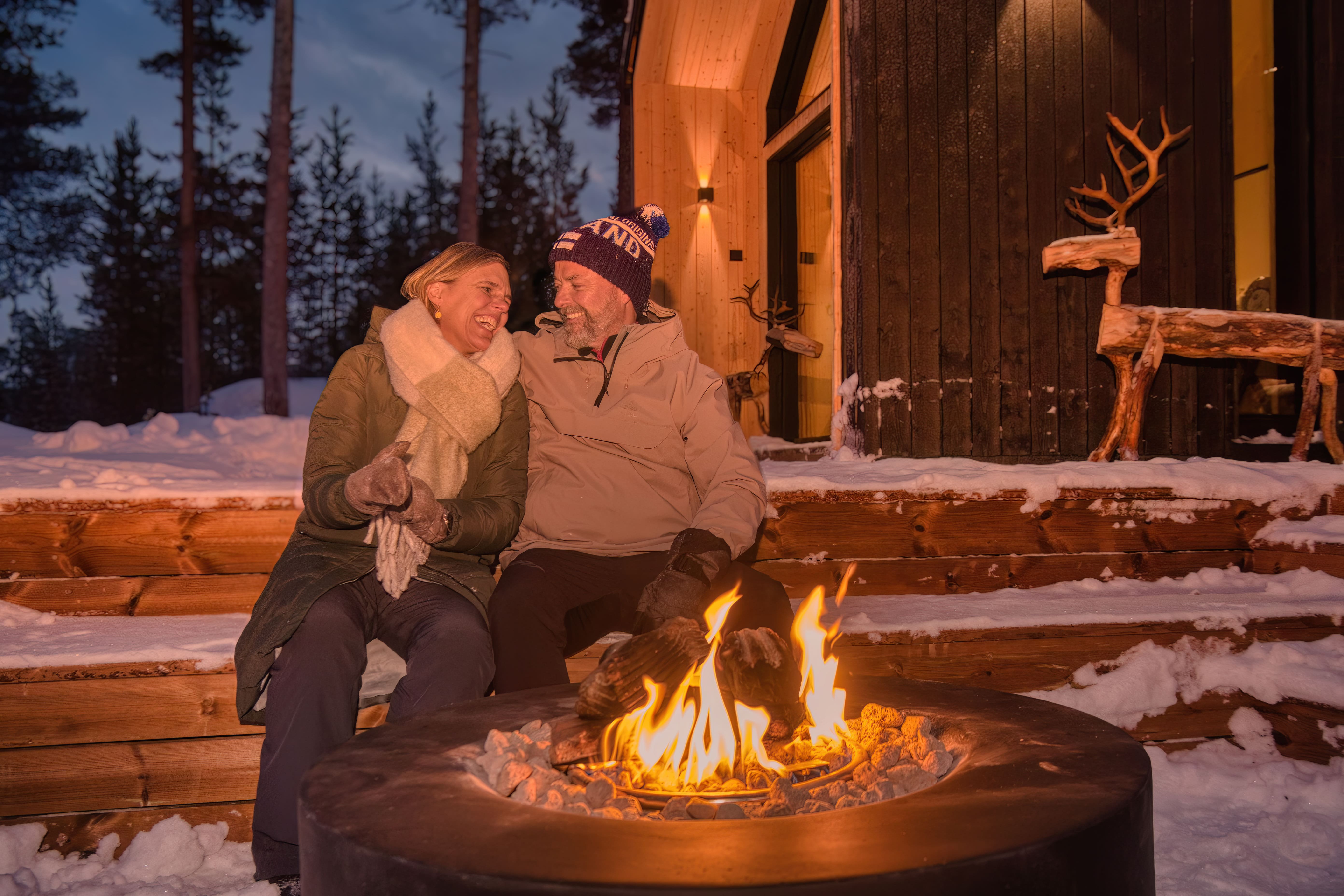 A laughing couple sits by a glowing fire pit on a snowy wooden platform, illuminated by warm orange light. They are dressed in winter coats and a beanie, with a dark blue evening sky and pine trees in the background. A modern wood cabin with wall lights and carved wooden reindeer figures stands behind them.