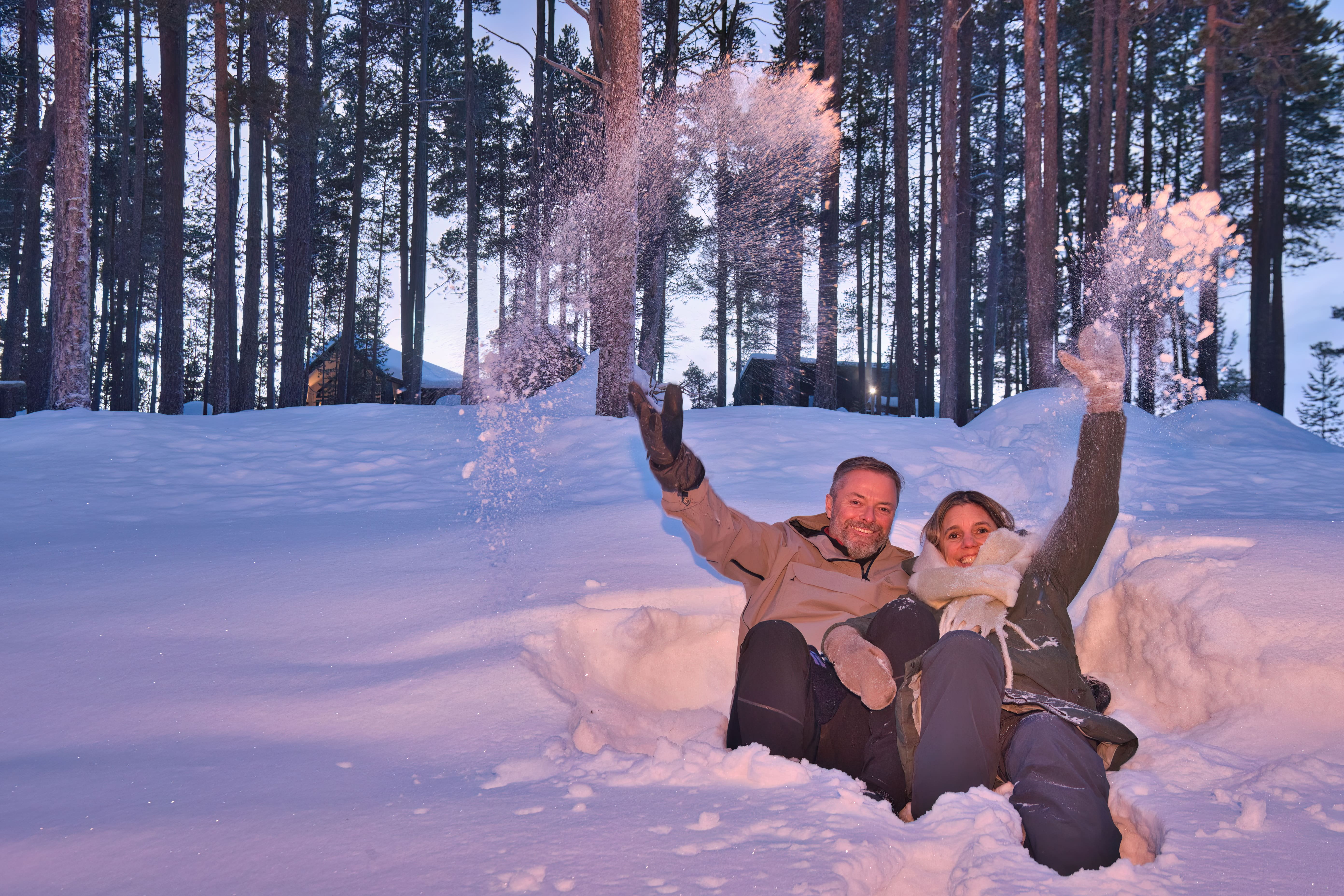 A smiling man and woman sit in a snowdrift in a winter forest, throwing snow into the air. The snow-covered landscape is illuminated with soft purple and pink light, while the airborne snow catches a warm, golden glow. Tall, dark pine trees frame the scene, with distant cabins visible in the background under a pale blue sky.