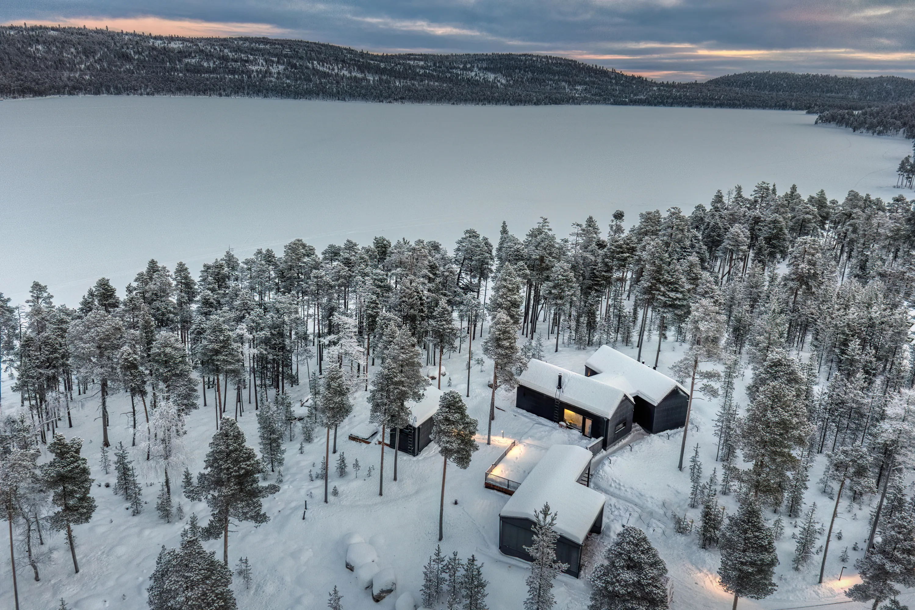 An aerial view reveals a serene winter landscape with a vast, frozen lake covered in white snow under a cloudy, blue-grey sky with soft orange hues. A dense forest of snow-laden pine trees fills the foreground, where several dark-colored, modern cabins with snow-covered roofs are nestled. Warm yellow light glows from some cabin windows and outdoor path lights, illuminating the pristine white snow.