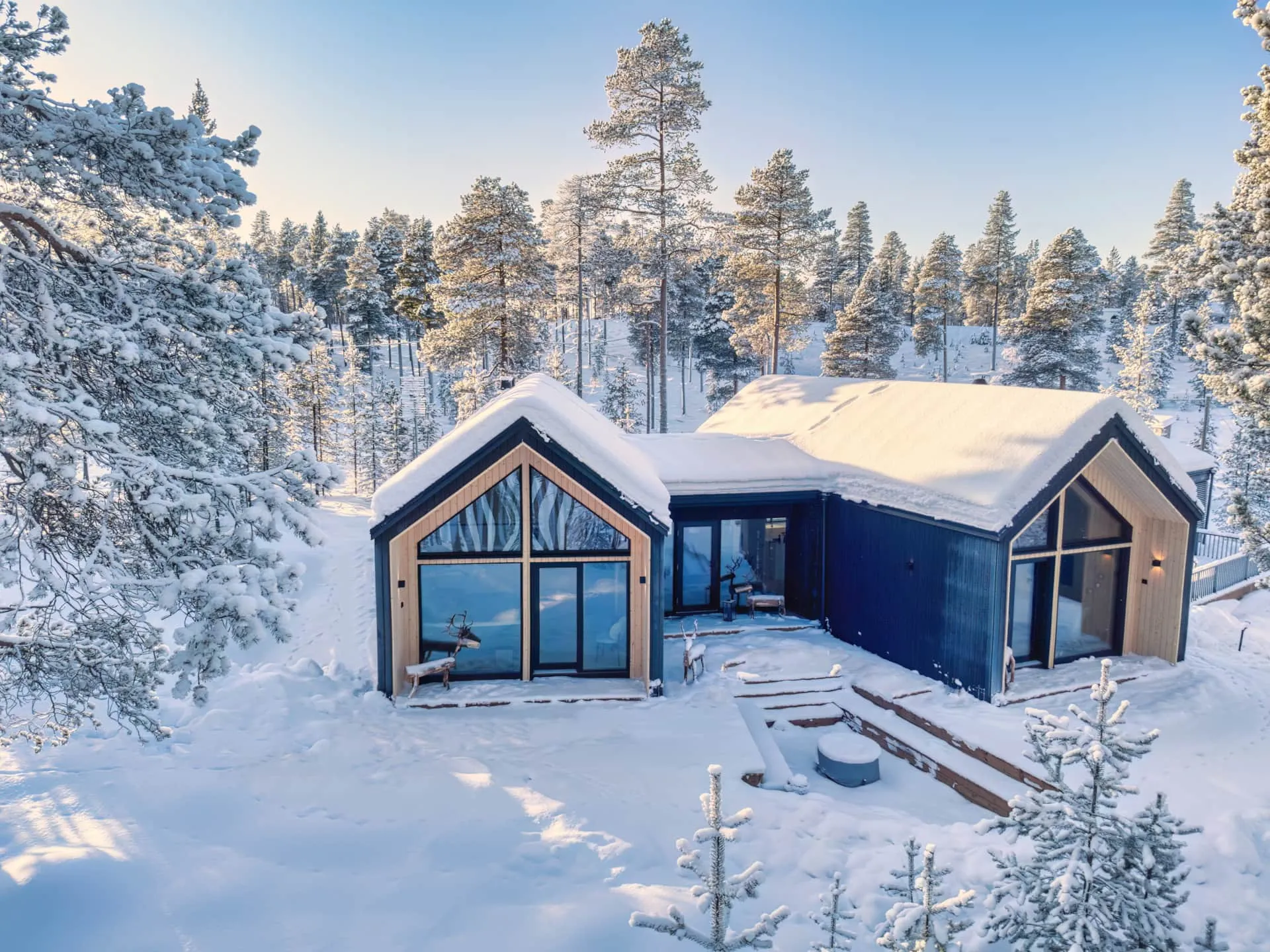 A modern dark blue and light wood house, its roofs blanketed in thick white snow, is nestled within a dense forest of snow-covered pine trees. Bright sunlight illuminates the winter scene, casting warm glows on the pristine snow and tree branches under a clear blue sky. Two wooden reindeer sculptures are positioned near the house's large glass windows and wooden steps.