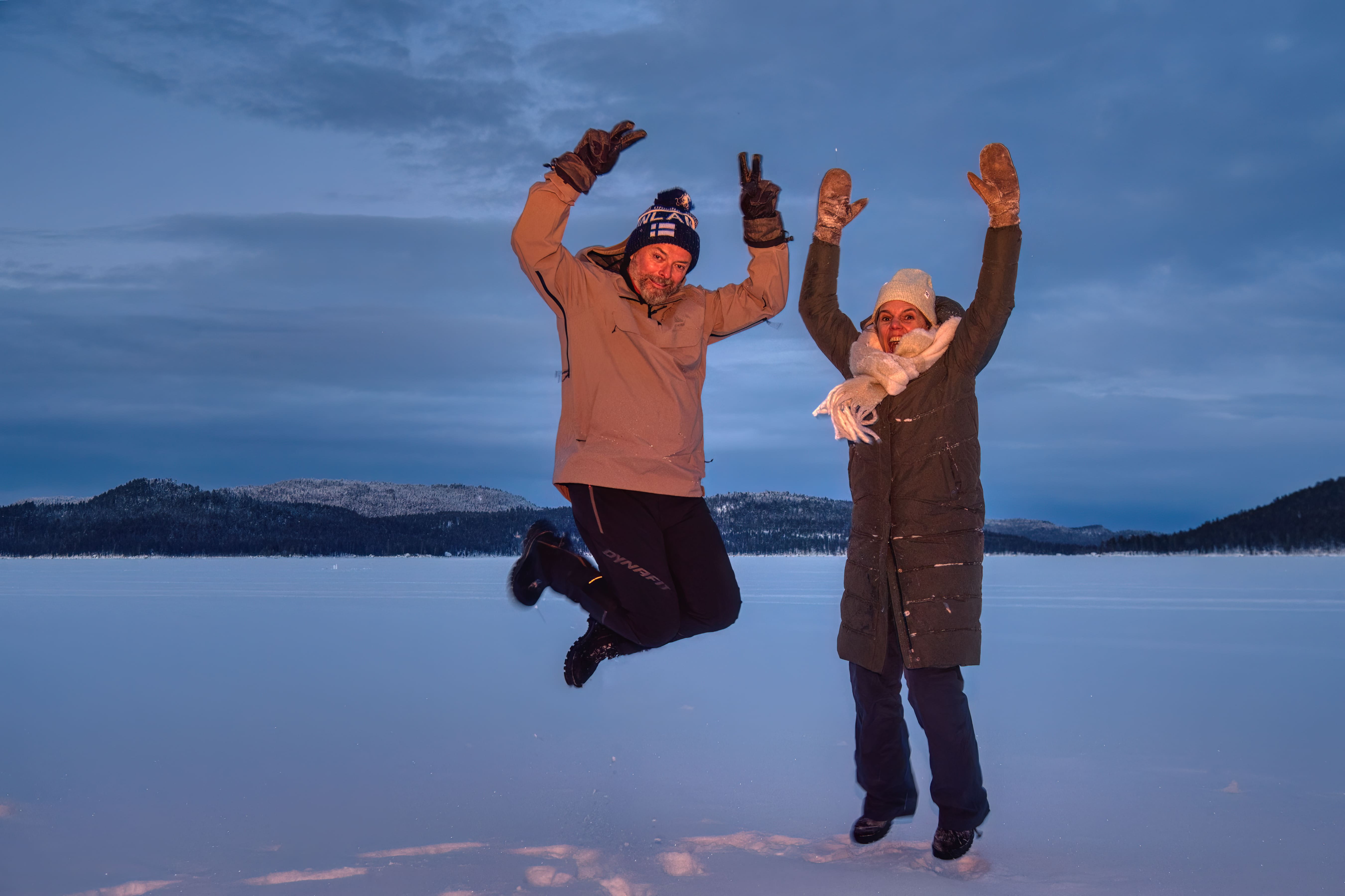 A man and a woman jump high in the air on a vast, snow-covered plain under a cloudy blue twilight sky. The man, on the left, wears a tan jacket and blue beanie, making a peace sign. The woman, on the right, wears a dark green coat, white scarf, and white beanie, smiling widely with arms raised. Dark, snow-dusted evergreen trees cover distant hills beneath the cool, muted light.