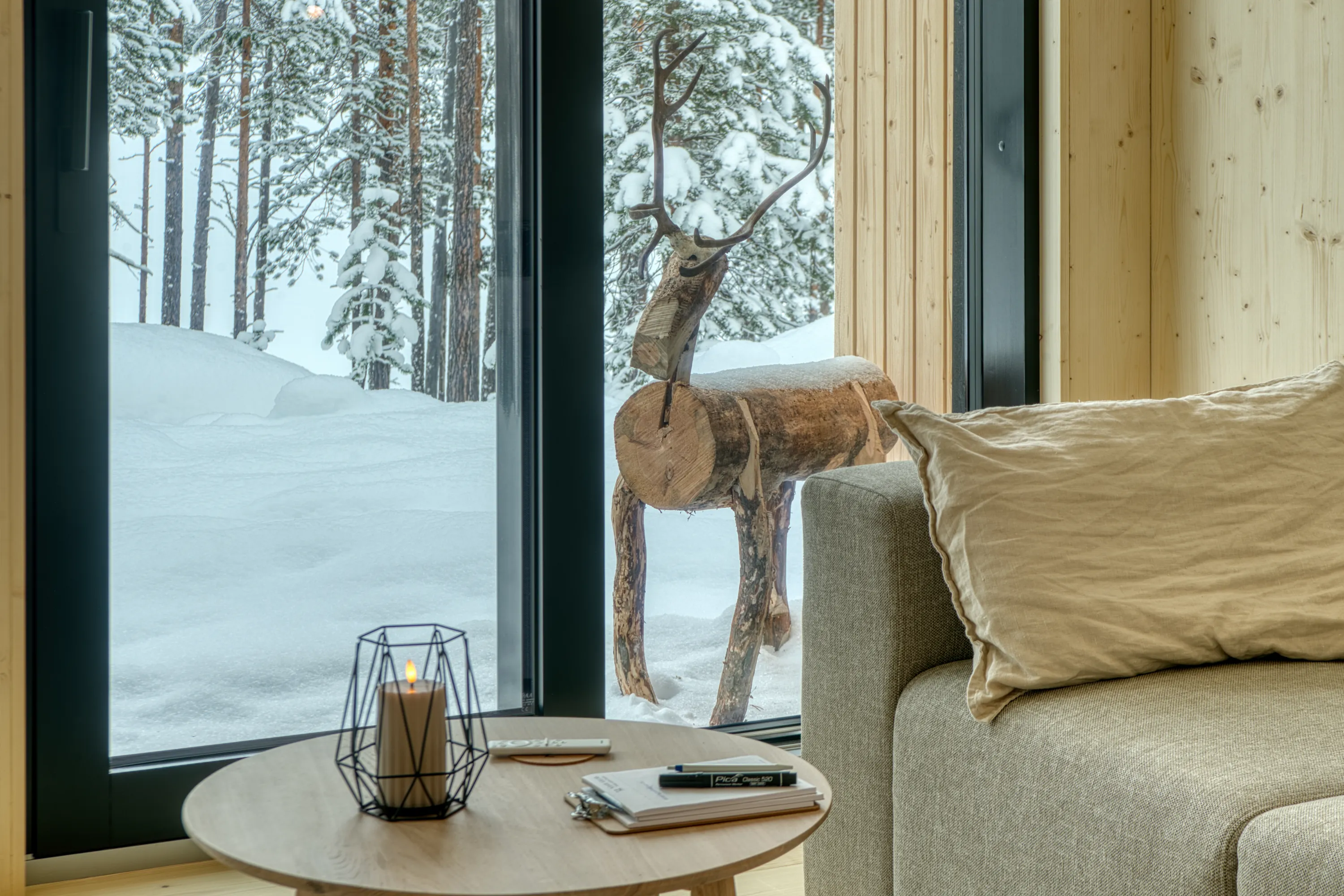 A cozy interior features a light wood table holding a black geometric candle holder with a glowing faux candle, a white remote, and papers, all positioned before a large window. Outside, a snow-covered forest with pine trees and a rustic wooden reindeer sculpture is visible. A light grey sofa with a beige cushion occupies the right, illuminated by bright natural light and a warm candle glow.