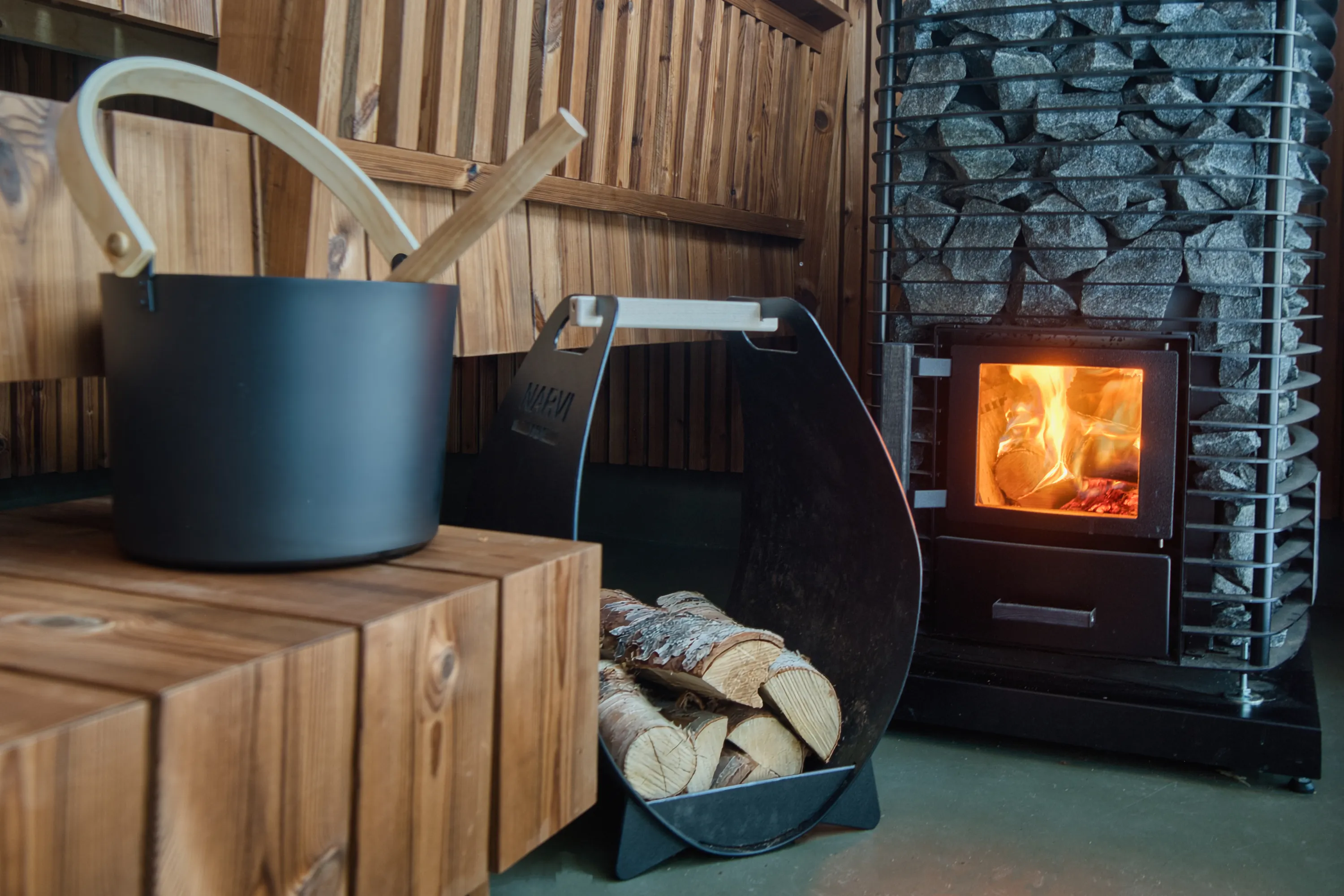 A warm sauna interior showcases a dark grey metal bucket with a light wooden handle and ladle on a light brown wooden bench. Adjacent is a black metal log holder filled with split firewood. On the right, a large sauna heater features a black metal cage packed with grey stones, and its firebox radiates bright orange and yellow flames from burning logs. The room has light brown wooden plank walls and a dark greenish-grey floor.