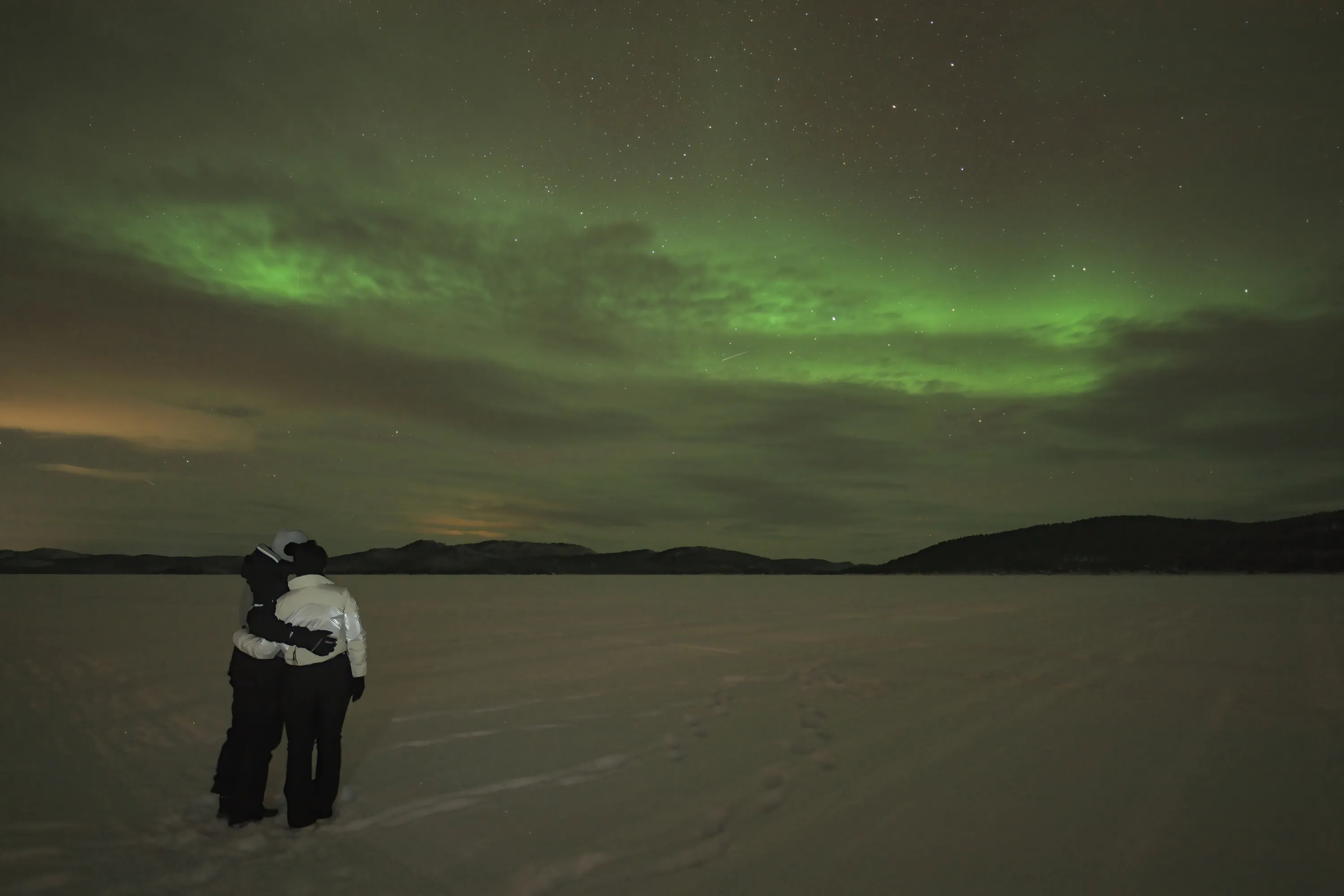Two people, seen from behind, stand embraced on a vast, snow-covered plain, looking up at a vibrant green aurora borealis. The night sky is filled with stars and faint clouds, with a subtle meteor trail visible. Dark mountains silhouette the horizon, and a soft orange glow appears on the far left, contrasting with the ethereal green light.