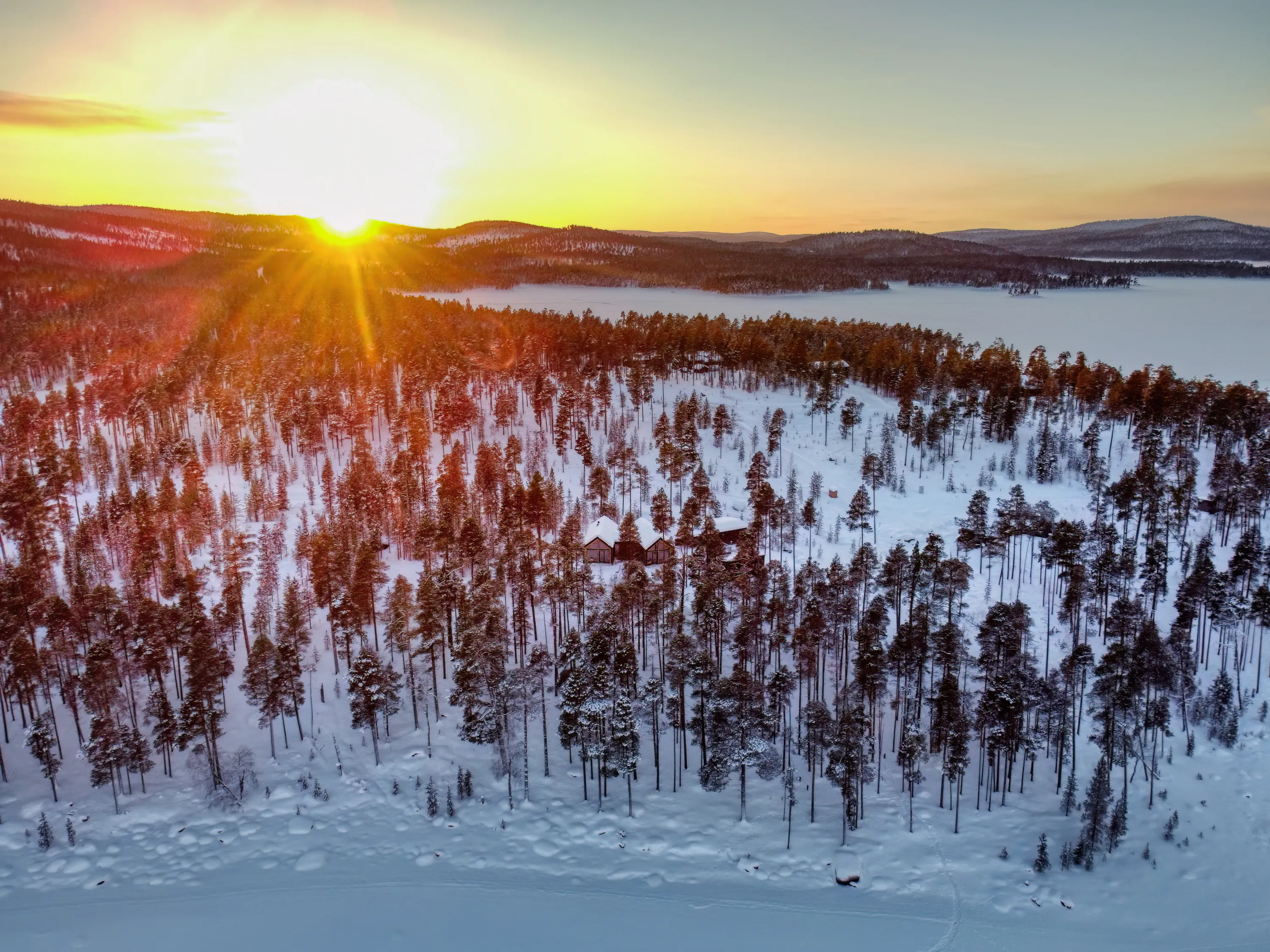 An aerial view captures a stunning winter sunset over a snow-covered landscape. The bright golden sun on the left casts warm rays across a dense forest of tall, snow-dusted evergreen trees, where several small cabins are nestled. A vast frozen lake, covered in white ice, stretches to the right, with distant, hazy mountains under a pale blue and orange sky.
