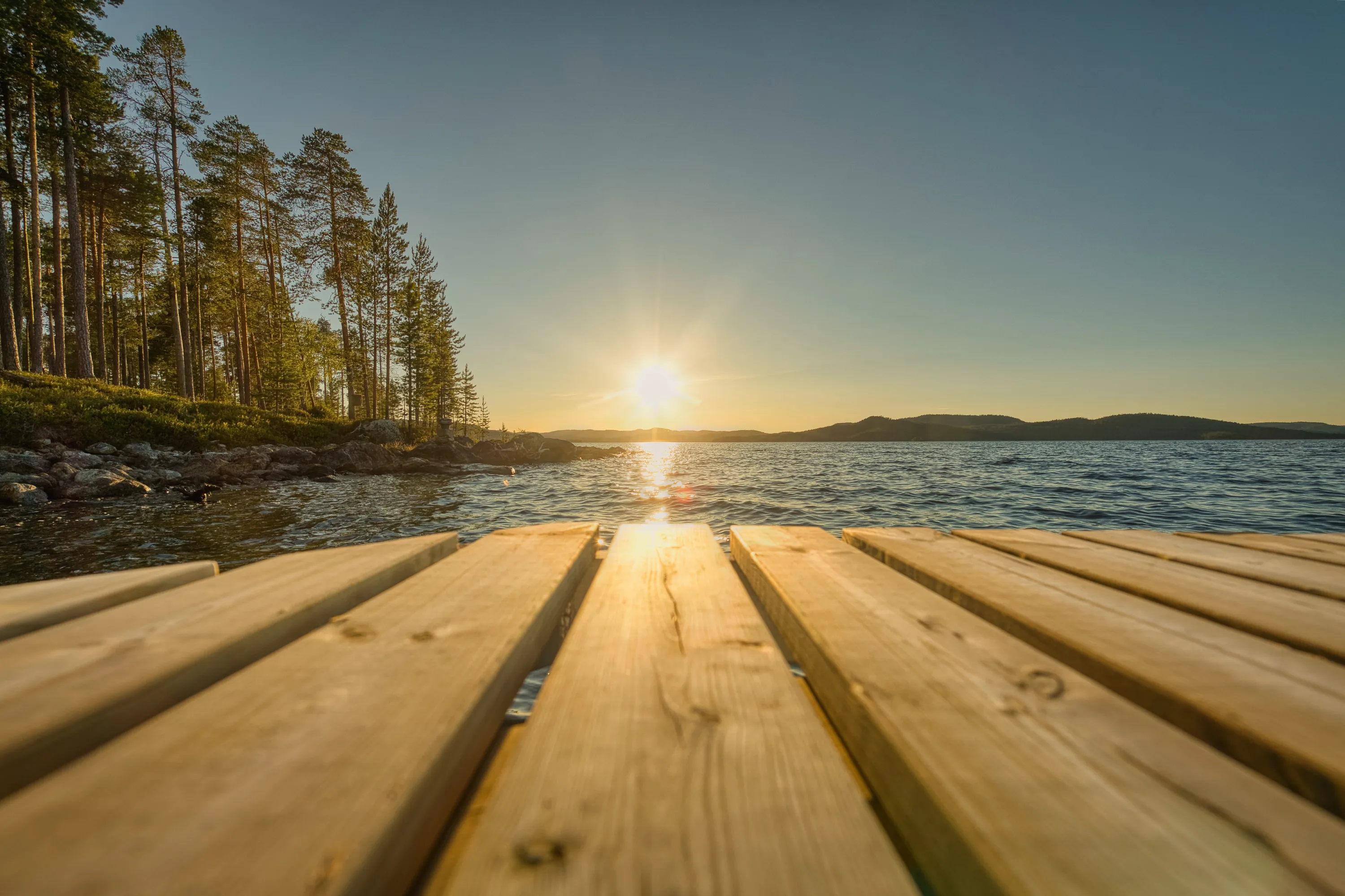 A low-angle view from a wooden pier extends over a calm lake at sunset. Golden sunlight illuminates the light brown planks in the foreground, reflecting brightly on the rippling dark blue water. To the left, a rocky shoreline with green foliage is backed by tall pine trees, bathed in warm light. Distant hills are silhouetted against the clear sky, which transitions from light blue to a radiant yellow-orange around the setting sun.