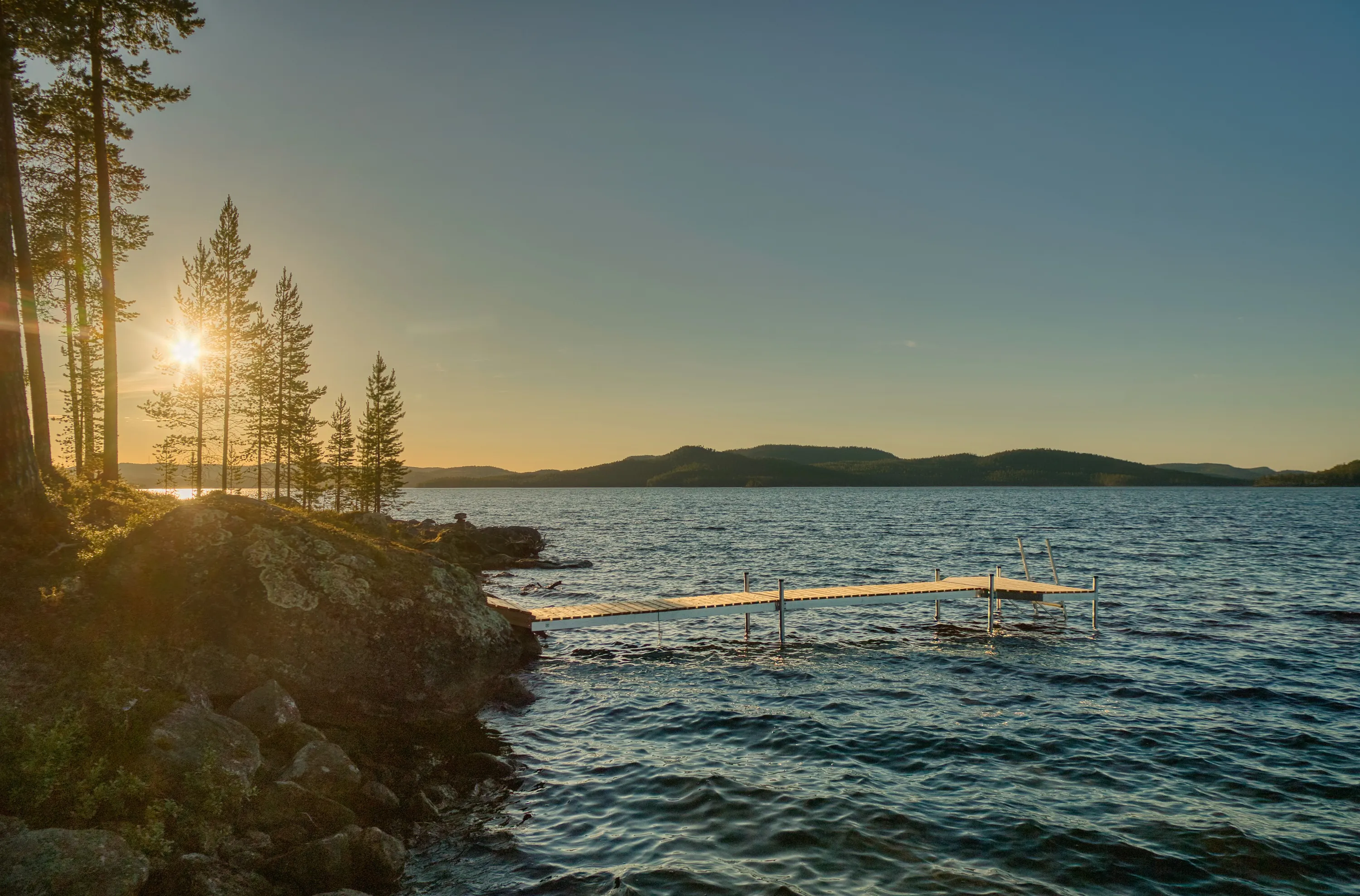 A serene lake scene at golden hour, with the sun low on the left, casting a bright lens flare and warm light through tall evergreen trees on a rocky bank. The sky transitions from a radiant orange-yellow near the horizon to a soft blue-grey overhead. A light-colored wooden dock with metal supports extends into the rippling, dark blue-green water, reflecting the sunlight. Distant forested hills are silhouetted against the horizon.
