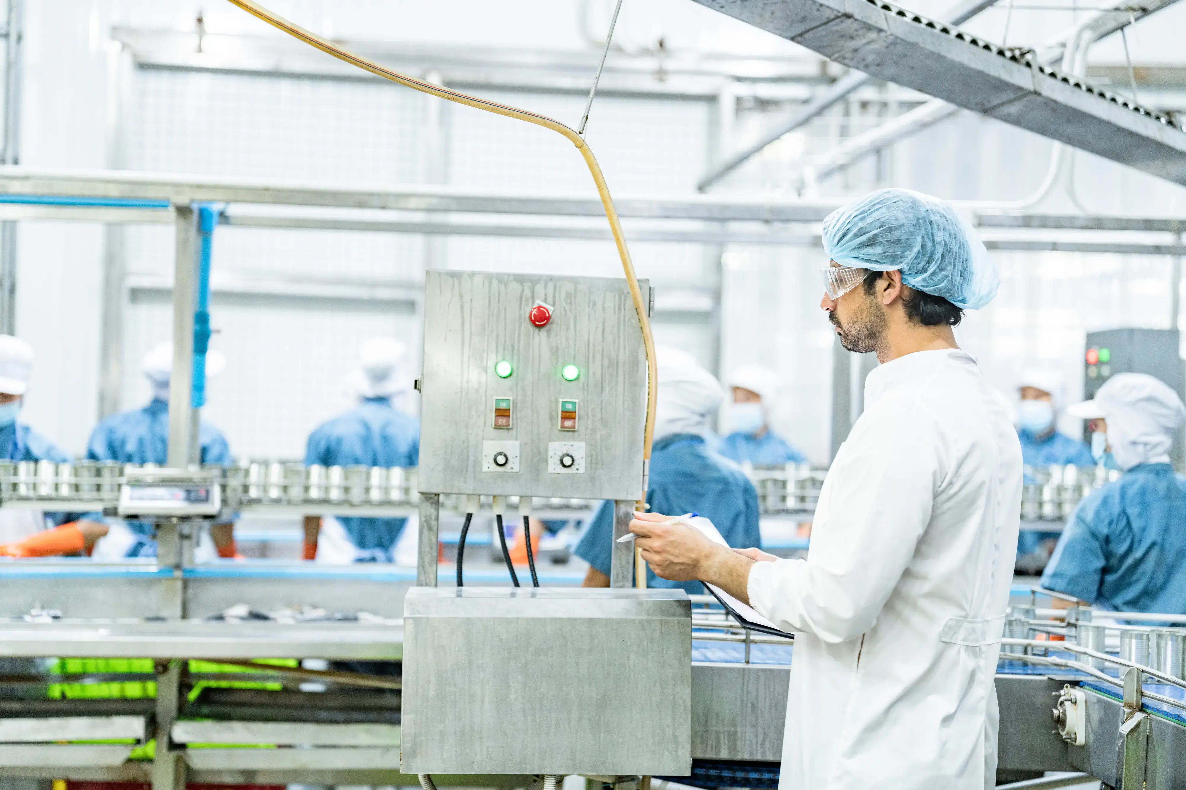 Factory worker in protective clothing and hairnet checking control panel in a food processing plant with other workers in the background.