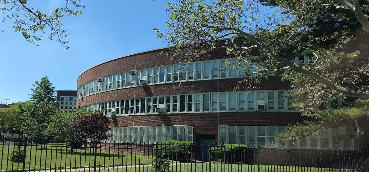 Curved three-story brick building with many windows behind a black metal fence, surrounded by green trees and grass under a clear blue sky.