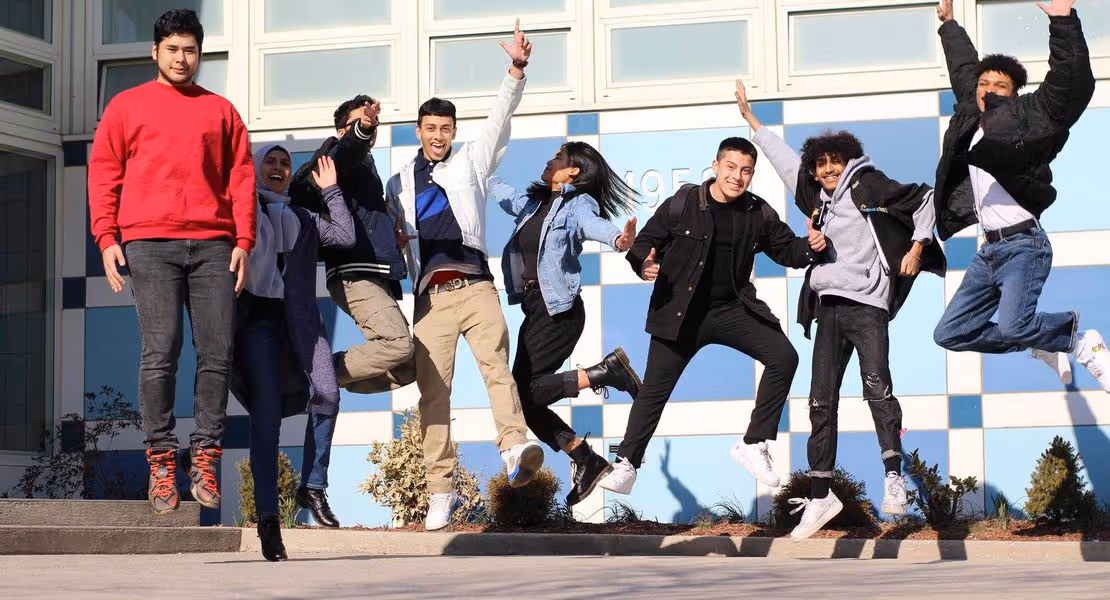 Group of eight diverse young people jumping in front of a blue and white checkered wall outdoors, smiling and celebrating.