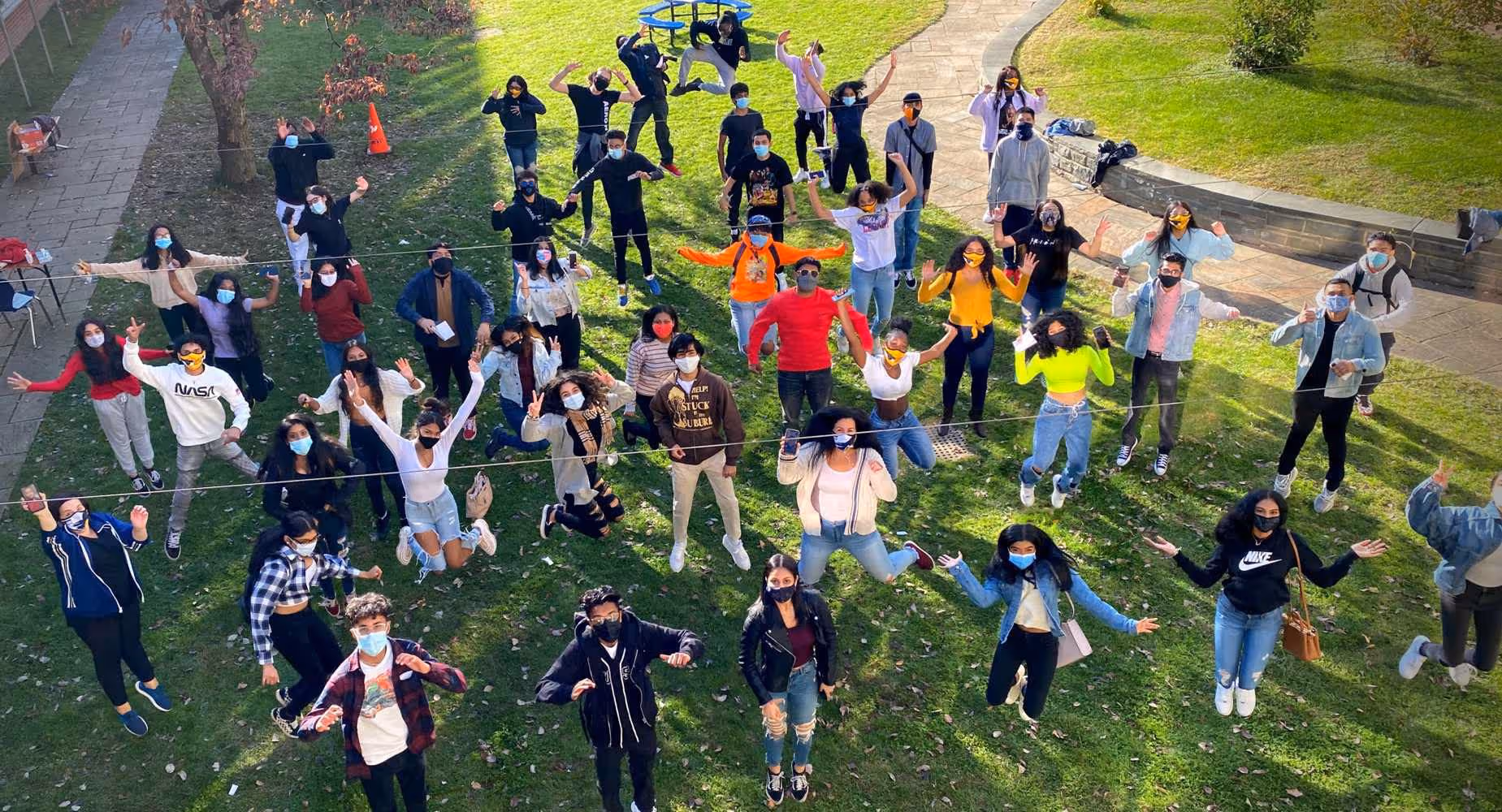 Group of young people wearing masks, standing and jumping on green grass in a park-like area.