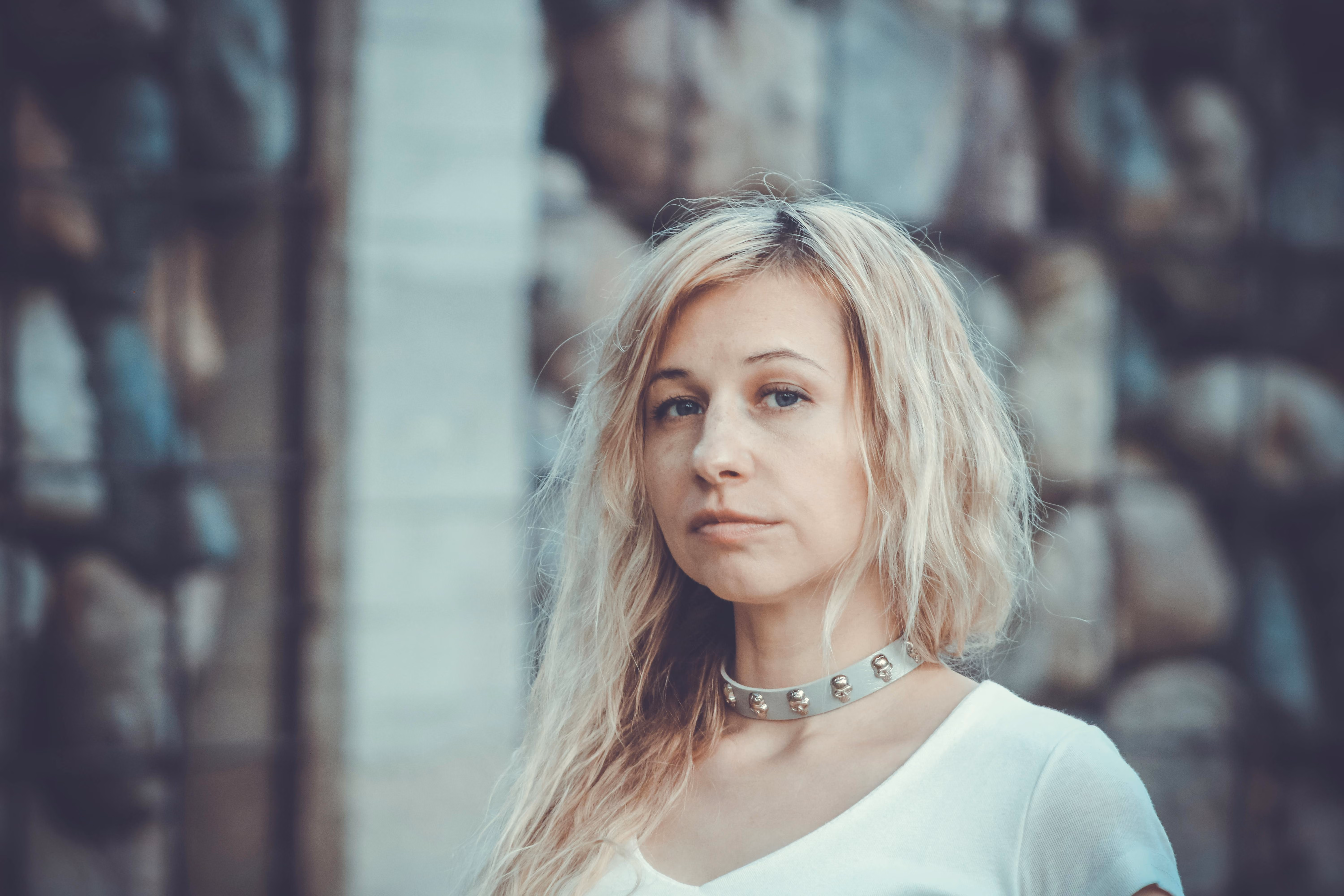Blonde woman with wavy hair wearing a white top and a choker necklace with skull designs against a blurred stone wall background.